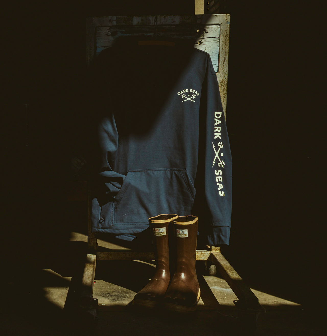 A dark navy blue pullover hoodie with 'DARK SEAS' printed in white on the sleeve and chest, displayed on a wooden stand next to a pair of brown xtratuf boots in a very dark setting.