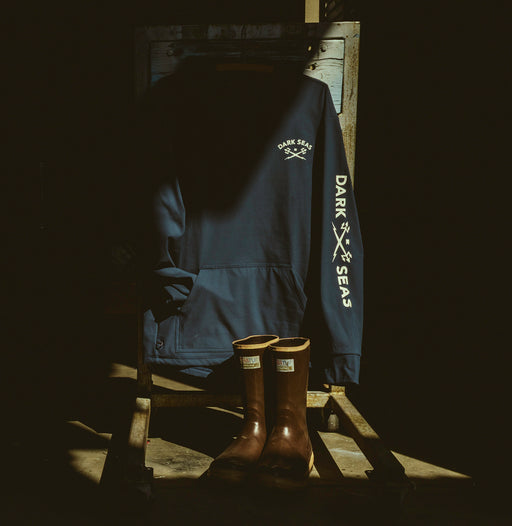 A dark navy blue pullover hoodie with 'DARK SEAS' printed in white on the sleeve and chest, displayed on a wooden stand next to a pair of brown xtratuf boots in a very dark setting.