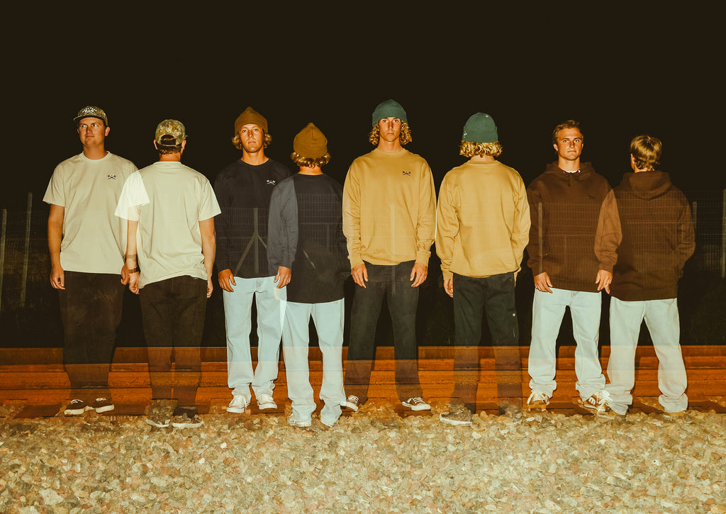 Eight men standing in a row outdoors at night, photographed from the back. They are wearing casual streetwear including t-shirts, hoodies, sweatshirts, jeans, and beanies.