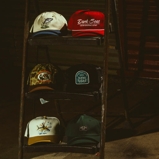 Four baseball caps displayed on a dark metal shelving unit in a dim indoor setting. The caps include a cream and blue cap, an orange-red cap with 'Dark Seas' text, a camouflage cap, a black patch cap, a cream and gold cap, and a dark green cap.