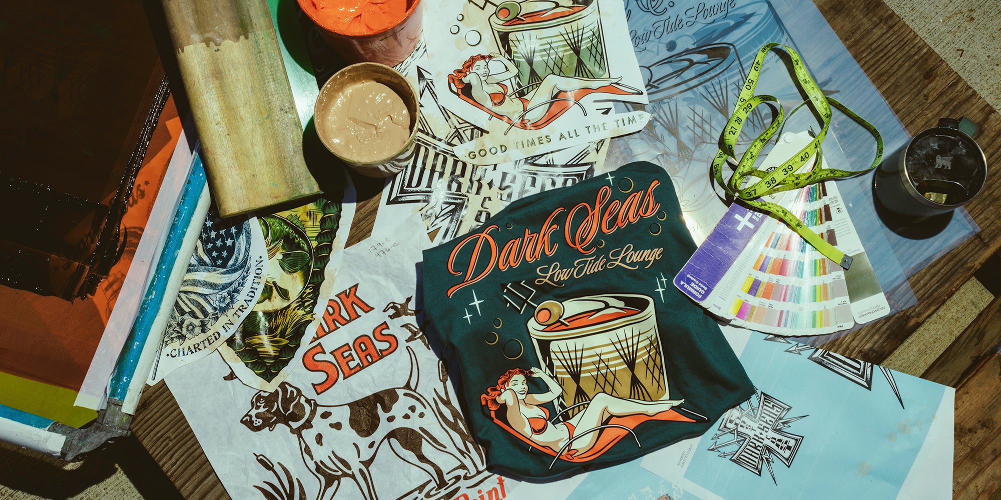 A black tote bag with a vintage-style 'Dark Seas' tattoo-style illustration and text lies on top of printed artwork sheets on a wooden table. Surrounding props include a green patterned lanyard, a small cup, a paint swatch fan, and other printed materials.