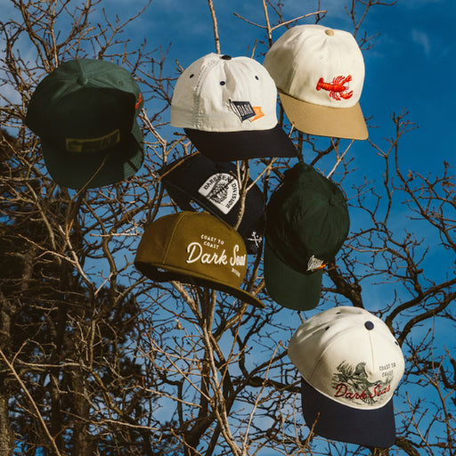 Assorted Dark Seas hats hanging form tree branches outdoors.