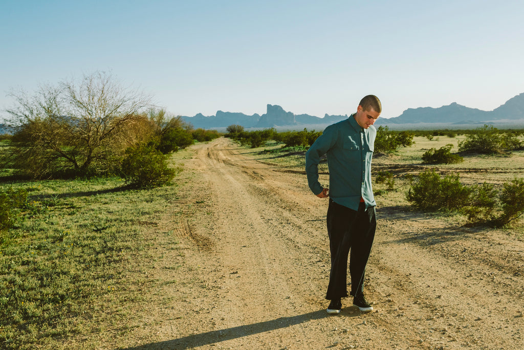 A man wearing a teal long-sleeve button-up shirt and black pants stands on a dirt road in a desert landscape with mountains in the background.