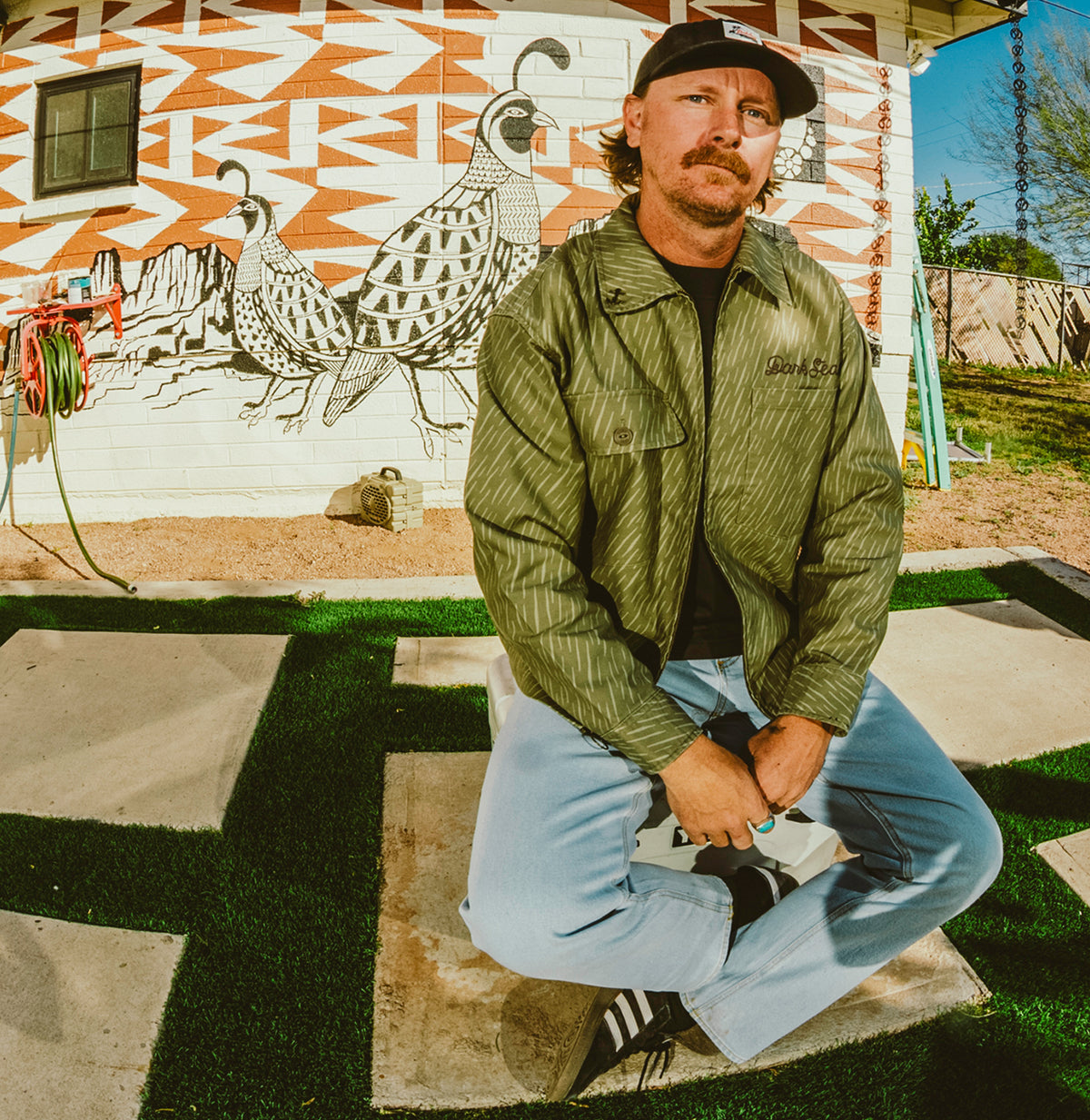Green rain camo pattern jacket on a man sitting in front of a wall mural with a quail and geometric shape layout.